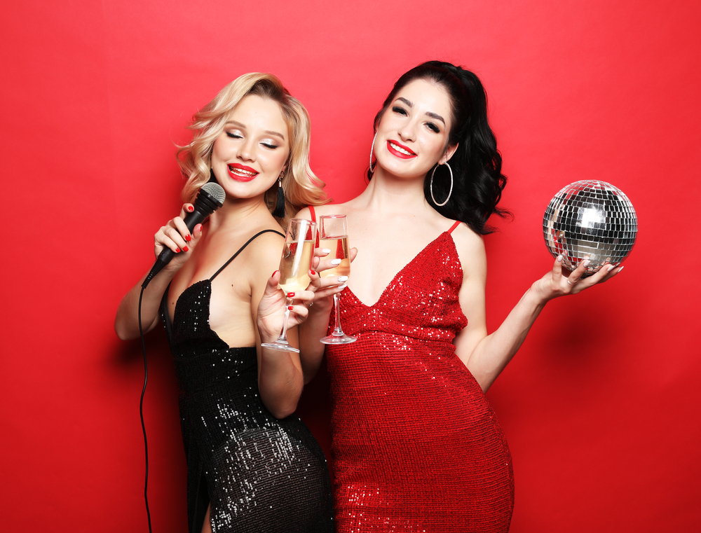 Two beautiful women, blonde and brunette wearing dresses, sing with a microphone, holding disco balls and glass of wine,